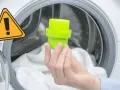 Main dans la machine à laver, tenant un bouchon vert pour le bac adoucissant. Un panneau d'avertissement jaune est visible.