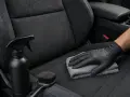 Main tenant, ganté, nettoie un siège de voiture en alcantara avec un chiffon. Un spray et une brosse sont à proximité, pour savoir comment nettoyer un canapé en alcantara.