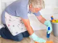 Un homme âgé, portant un tablier à étoiles, utilise une astuce de grand-mère pour nettoyer le lino avec un spray et un chiffon.