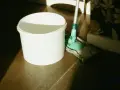 Seau blanc et serpillère verte prêts pour un nettoyage de sol maison. La lumière du soleil éclaire la scène.