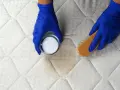 Mains gantées enfilées, on s'apprête à enlever une tache de sang sur un matelas avec une poudre blanche et une brosse.