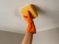 Main tenant nettoie le plafond taché avec une éponge jaune. Le sol sale n'est plus un problème.