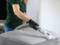 Personne avec des gants noirs utilise un aspirateur pour nettoyer un canapé en tissu. Une solution de vinaigre blanc et bicarbonate peut aider à nettoyer le canapé.