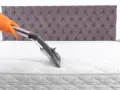Main tenant, gant orange, utilise un aspirateur pour comment nettoyer un matelas à sec. Le matelas est blanc, avec un couvre-lit capitonné gris en arrière-plan.