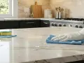 Main dans un gant blanc essuyant un comptoir en marbre avec un chiffon bleu. Des produits de nettoyage et des éponges sont visibles. Un bon traitement pierre pour une cuisine éclatante.