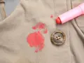 Rouge à lèvres sur polyester : une tache rouge vif sur un tissu beige, à côté d'un bouton marron et du tube de rouge à lèvres rose. Comment enlever cette tache ?