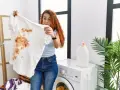 Femme surprise devant un t-shirt blanc taché de marron, prête à détacher le linge blanc. Machine à laver et lessive à proximité.