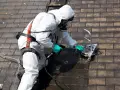 Nettoyage d'un toit en fibro ciment avec un appareil de nettoyage sous pression, l'opérateur porte une combinaison de protection et un masque.