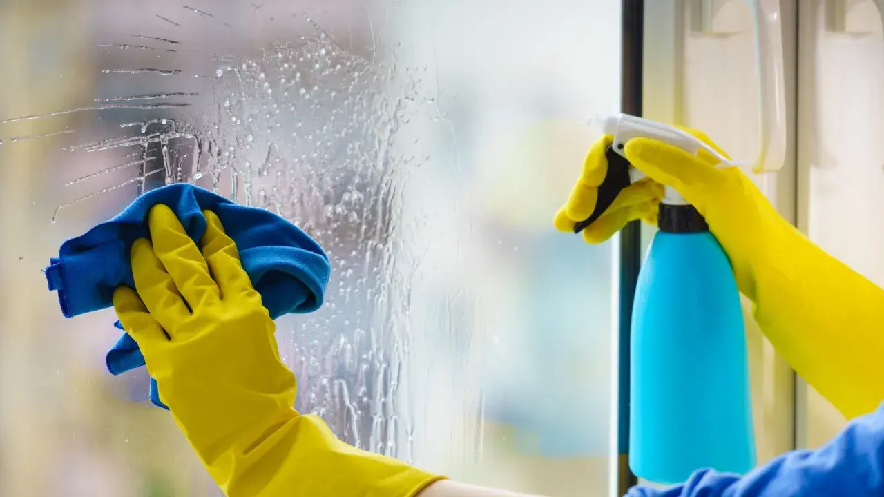 Mains gantées de jaune effectuent le nettoyage vitre avec un spray bleu et un chiffon bleu.