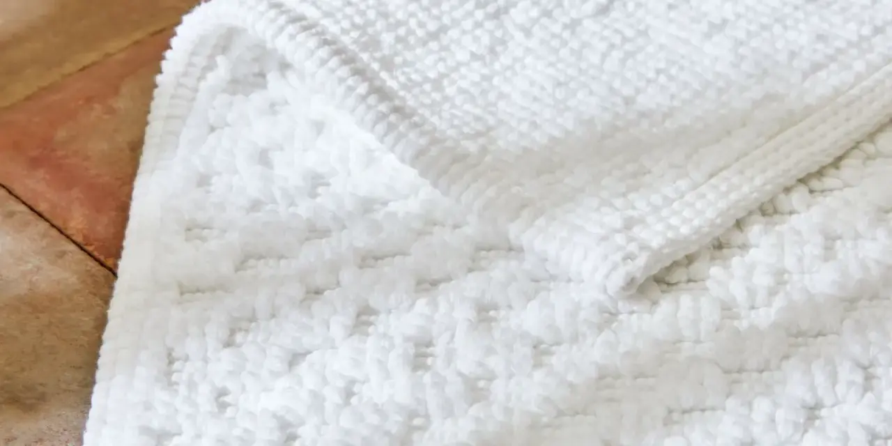 Un tapis de bain blanc moelleux, avec un motif texturé, est posé sur un sol en terre cuite. Il est temps de se demander a combien laver un tapis de bain pour qu'il reste ainsi.