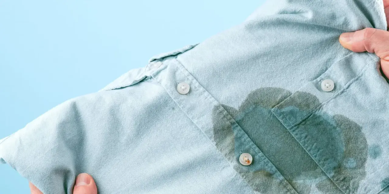 Main d'une personne tenant une chemise bleu pâle avec une grande tache d'huile sur le devant, prête à enlever une tache d'huile avec du bicarbonate.