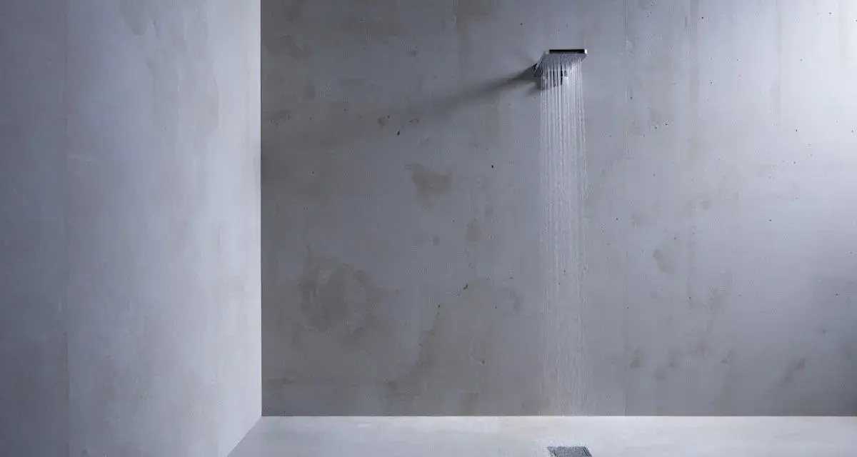 Douche minimaliste avec pommeau carré, eau tombant sur un sol en béton ciré sur galet. L'ambiance est épurée et moderne.