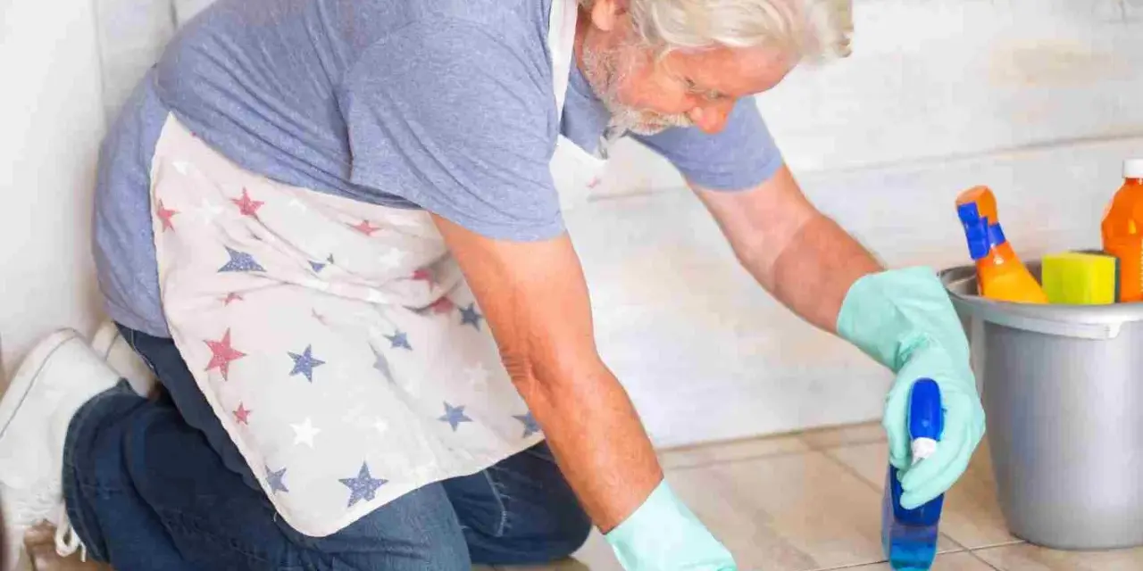 Un homme âgé, portant un tablier à étoiles, utilise une astuce de grand-mère pour nettoyer le lino avec un spray et un chiffon.