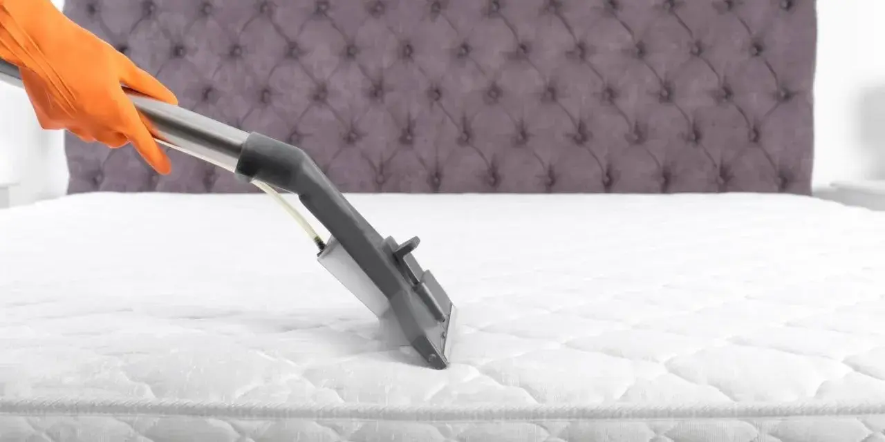 Main tenant, gant orange, utilise un aspirateur pour comment nettoyer un matelas à sec. Le matelas est blanc, avec un couvre-lit capitonné gris en arrière-plan.