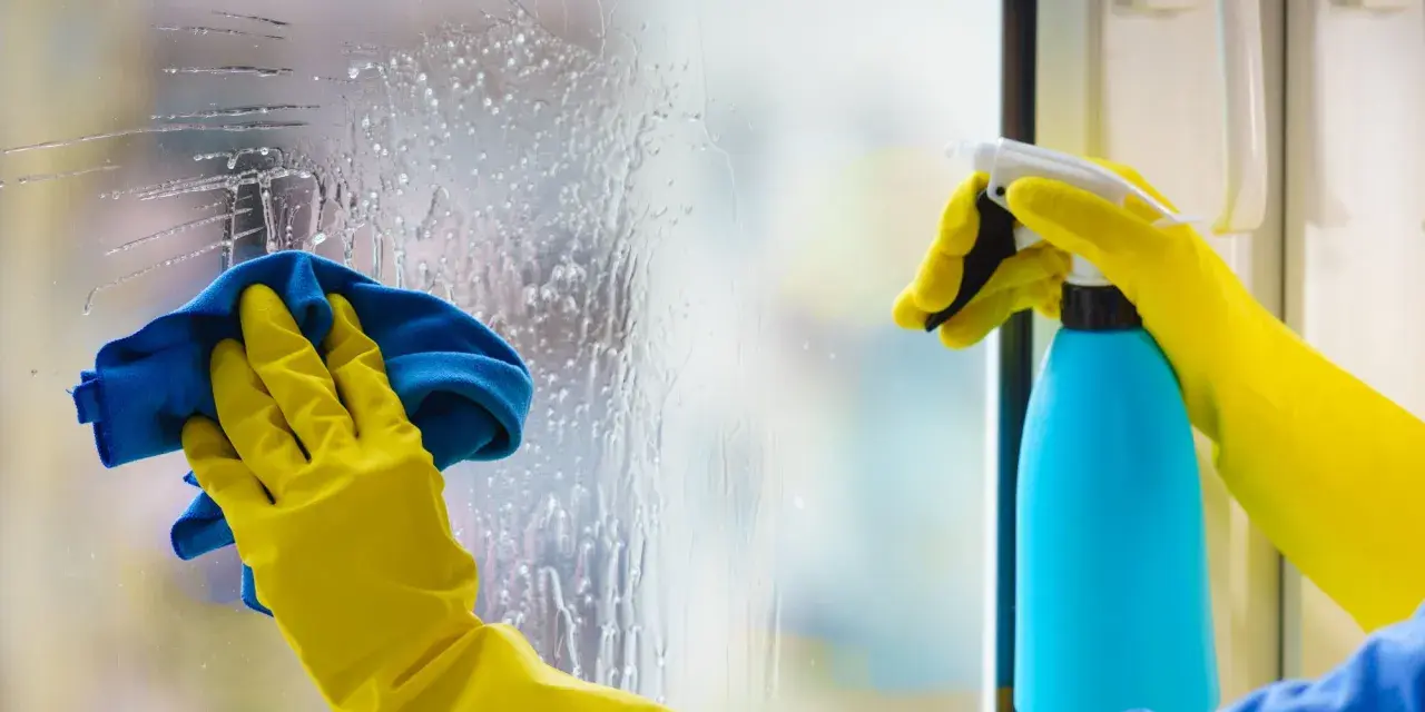 Mains gantées de jaune effectuent le nettoyage vitre avec un spray bleu et un chiffon bleu.