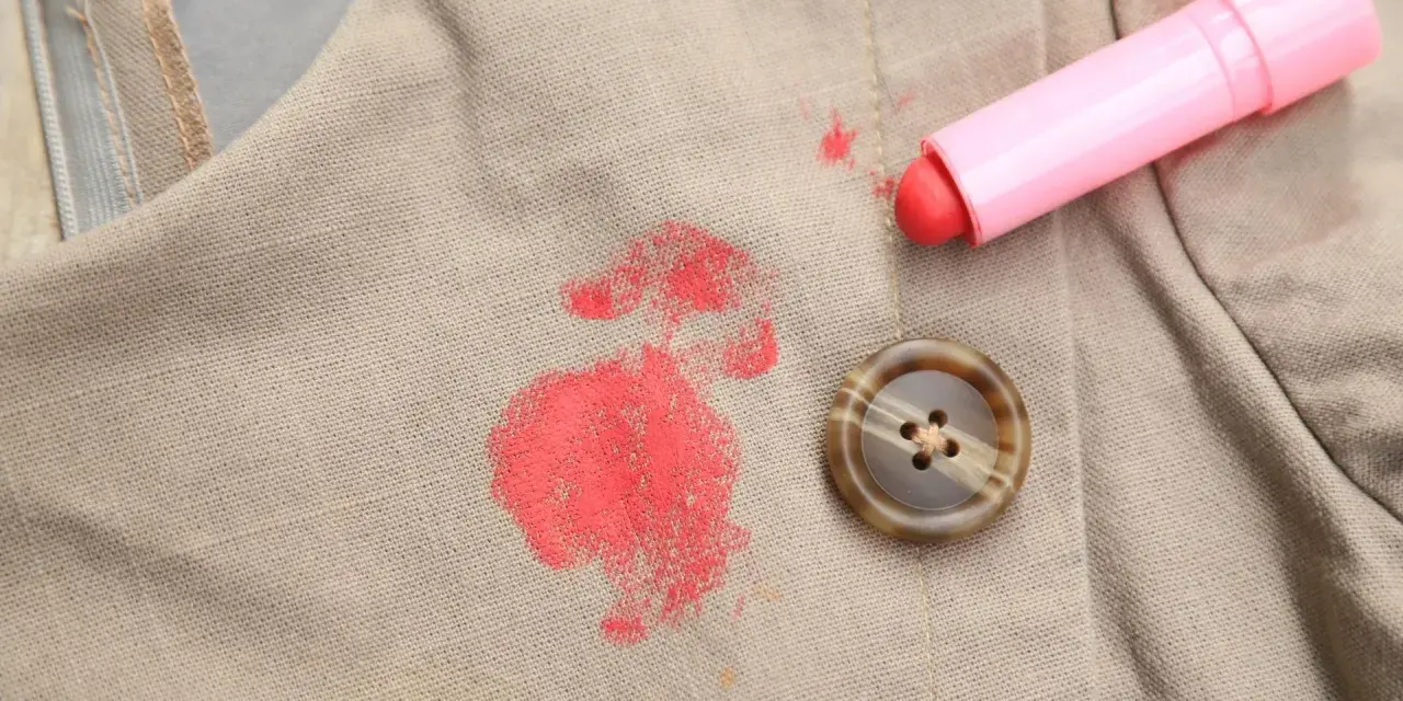 Rouge à lèvres sur polyester : une tache rouge vif sur un tissu beige, à côté d'un bouton marron et du tube de rouge à lèvres rose. Comment enlever cette tache ?