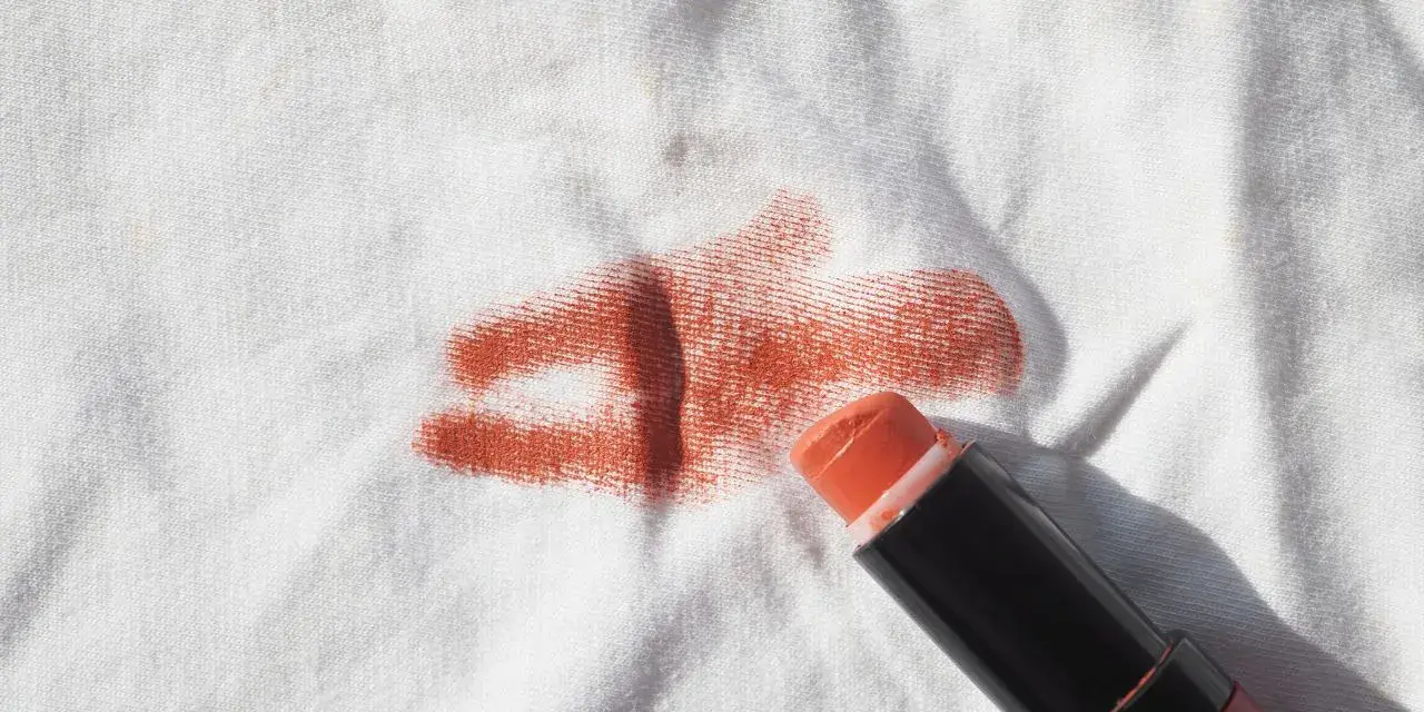 Un bâton de rouge à lèvres orange est posé sur un tissu blanc, laissant une tache rouge à lèvre.