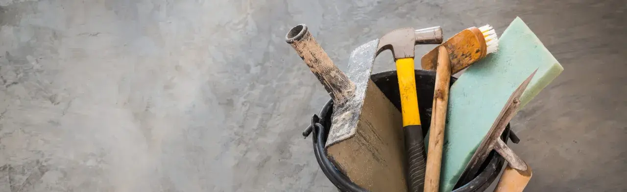 Outils pour nettoyer du ciment noirci : marteau, truelle, brosse et éponge sur fond de béton.