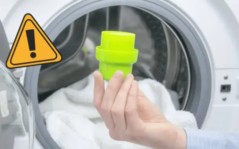 Main dans la machine à laver, tenant un bouchon vert pour le bac adoucissant. Un panneau d'avertissement jaune est visible.