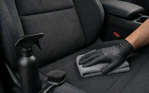 Main tenant, ganté, nettoie un siège de voiture en alcantara avec un chiffon. Un spray et une brosse sont à proximité, pour savoir comment nettoyer un canapé en alcantara.
