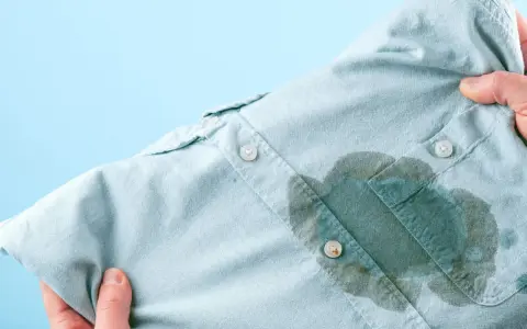 Main d'une personne tenant une chemise bleu pâle avec une grande tache d'huile sur le devant, prête à enlever une tache d'huile avec du bicarbonate.