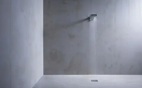 Douche minimaliste avec pommeau carré, eau tombant sur un sol en béton ciré sur galet. L'ambiance est épurée et moderne.