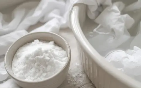 Un bol de bicarbonate de soude à côté d'un plat rempli d'eau et de linge blanc. Idéal pour éliminer les taches jaunes.
