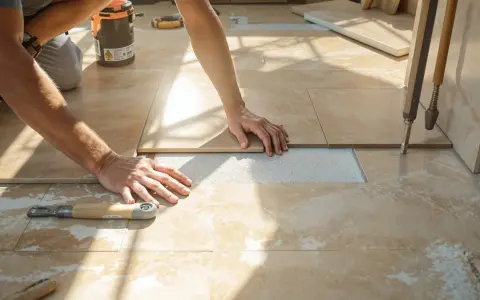 Pose d'un carrelage imitation travertin. Mains ajustant une dalle sur le lit de mortier. Outil de carreleur à proximité.