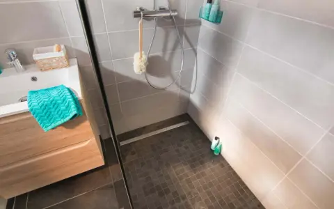 Sol de douche en galets sombres, contrastant avec le carrelage clair des murs. Une paroi vitrée sépare l'espace.