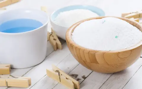 Bol en bois rempli de lessive en poudre maison, à côté d'une tasse de liquide bleu et de pinces à linge en bois.