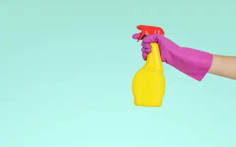 Main tenant, une main gantée de rose tient un flacon jaune prêt à nettoyer le sol en vinyle avec du vinaigre blanc.