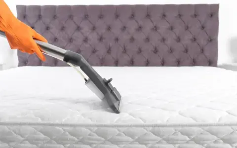 Main tenant, gant orange, utilise un aspirateur pour comment nettoyer un matelas à sec. Le matelas est blanc, avec un couvre-lit capitonné gris en arrière-plan.