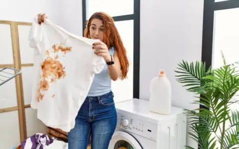 Femme surprise devant un t-shirt blanc taché de marron, prête à détacher le linge blanc. Machine à laver et lessive à proximité.