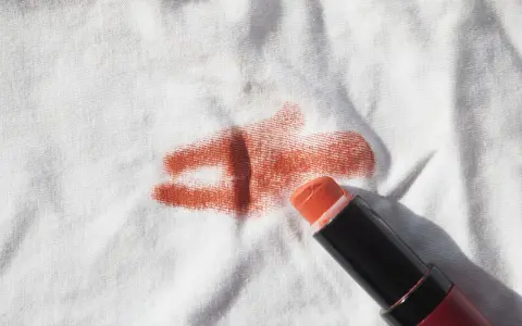 Un bâton de rouge à lèvres orange est posé sur un tissu blanc, laissant une tache rouge à lèvre.