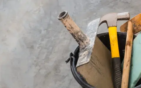 Outils pour nettoyer du ciment noirci : marteau, truelle, brosse et éponge sur fond de béton.