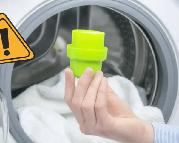 Main dans la machine à laver, tenant un bouchon vert pour le bac adoucissant. Un panneau d'avertissement jaune est visible.