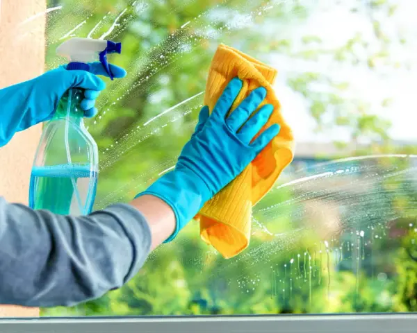Mains gantées de bleu pulvérisent un produit pour nettoyer les vitres sans traces, puis essuient avec un chiffon jaune.