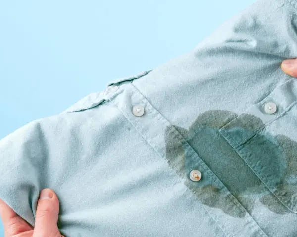 Main d'une personne tenant une chemise bleu pâle avec une grande tache d'huile sur le devant, prête à enlever une tache d'huile avec du bicarbonate.
