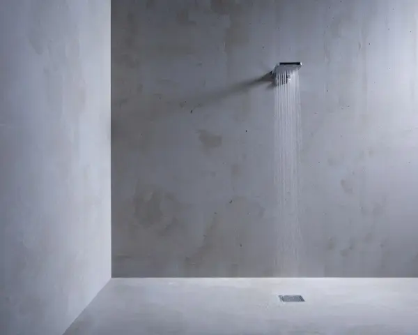 Douche minimaliste avec pommeau carré, eau tombant sur un sol en béton ciré sur galet. L'ambiance est épurée et moderne.