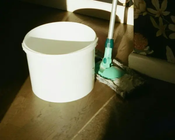 Seau blanc et serpillère verte prêts pour un nettoyage de sol maison. La lumière du soleil éclaire la scène.