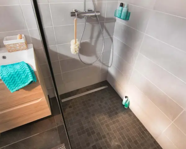 Sol de douche en galets sombres, contrastant avec le carrelage clair des murs. Une paroi vitrée sépare l'espace.