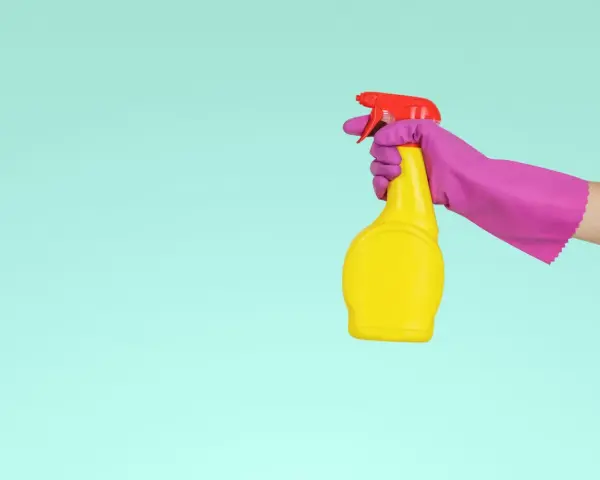 Main tenant, une main gantée de rose tient un flacon jaune prêt à nettoyer le sol en vinyle avec du vinaigre blanc.