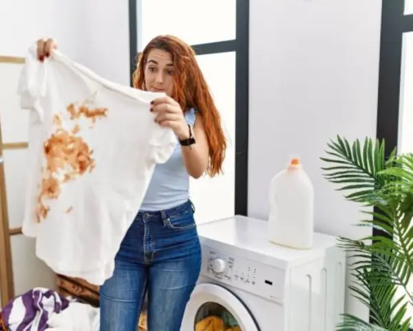 Femme surprise devant un t-shirt blanc taché de marron, prête à détacher le linge blanc. Machine à laver et lessive à proximité.