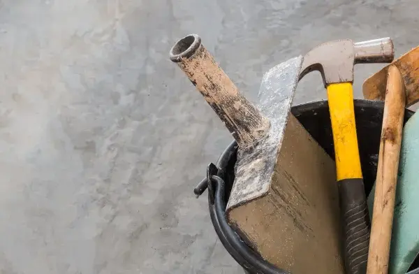 Outils pour nettoyer du ciment noirci : marteau, truelle, brosse et éponge sur fond de béton.