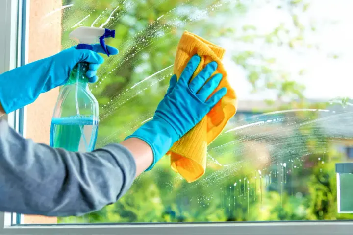 Mains gantées de bleu pulvérisent un produit pour nettoyer les vitres sans traces, puis essuient avec un chiffon jaune.