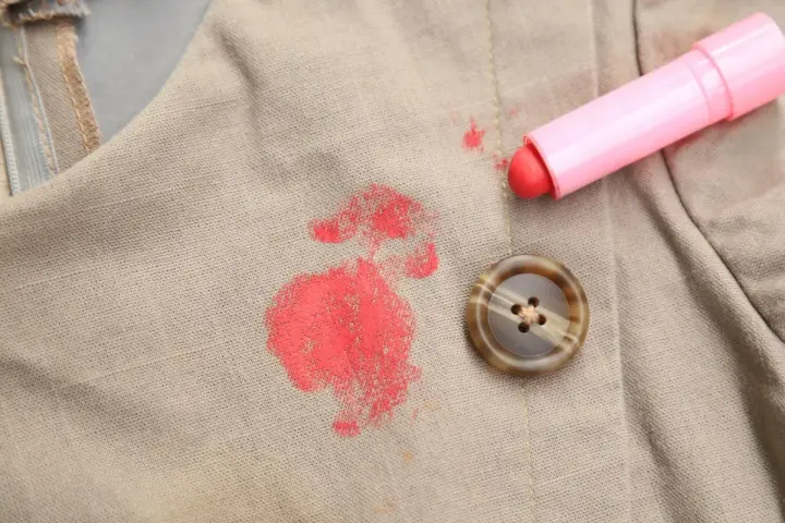Rouge à lèvres sur polyester : une tache rouge vif sur un tissu beige, à côté d'un bouton marron et du tube de rouge à lèvres rose. Comment enlever cette tache ?