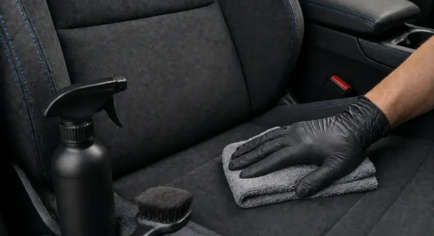 Main tenant, ganté, nettoie un siège de voiture en alcantara avec un chiffon. Un spray et une brosse sont à proximité, pour savoir comment nettoyer un canapé en alcantara.