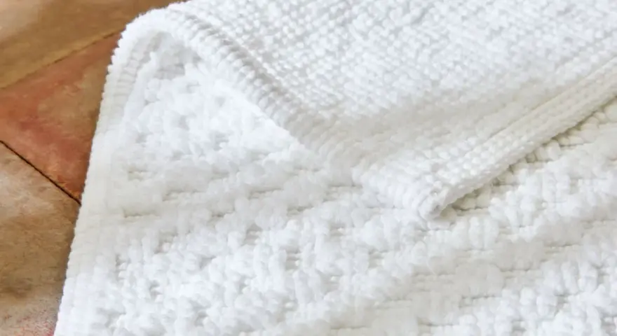 Un tapis de bain blanc moelleux, avec un motif texturé, est posé sur un sol en terre cuite. Il est temps de se demander a combien laver un tapis de bain pour qu'il reste ainsi.