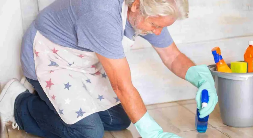 Un homme âgé, portant un tablier à étoiles, utilise une astuce de grand-mère pour nettoyer le lino avec un spray et un chiffon.