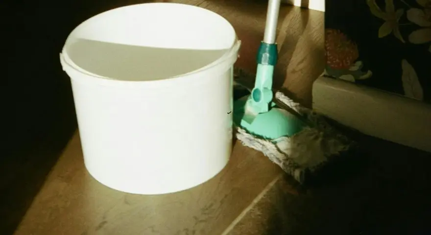 Seau blanc et serpillère verte prêts pour un nettoyage de sol maison. La lumière du soleil éclaire la scène.